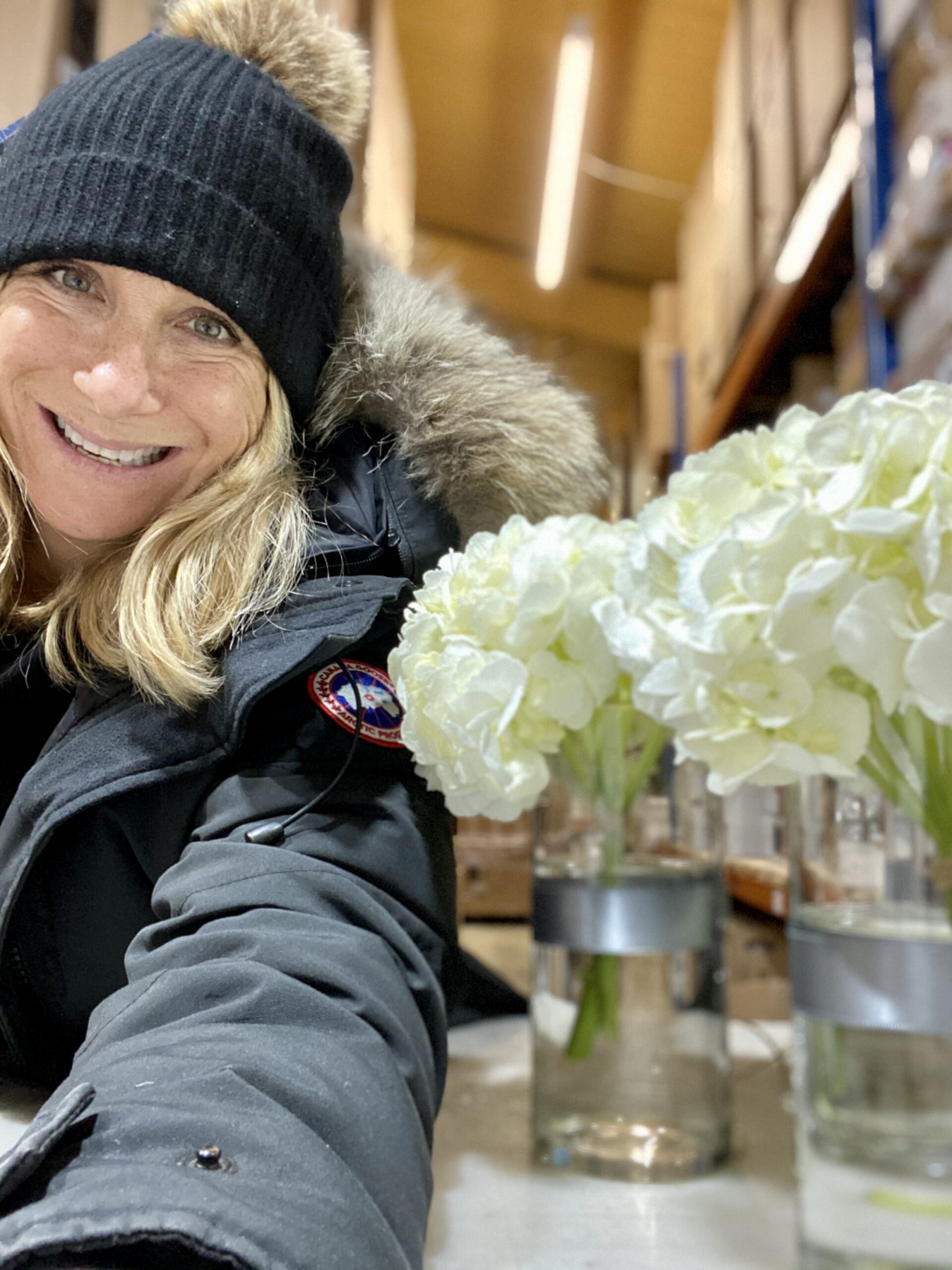blonde woman smiling next to a pot of flowers. she is wearing a black hat.